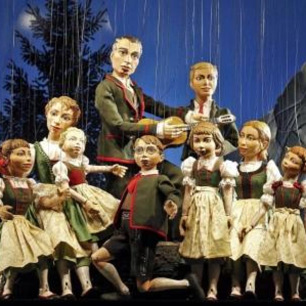 Puppen in traditioneller österreichischer Kleidung treten auf der Bühne vor einer Bergkulisse auf.