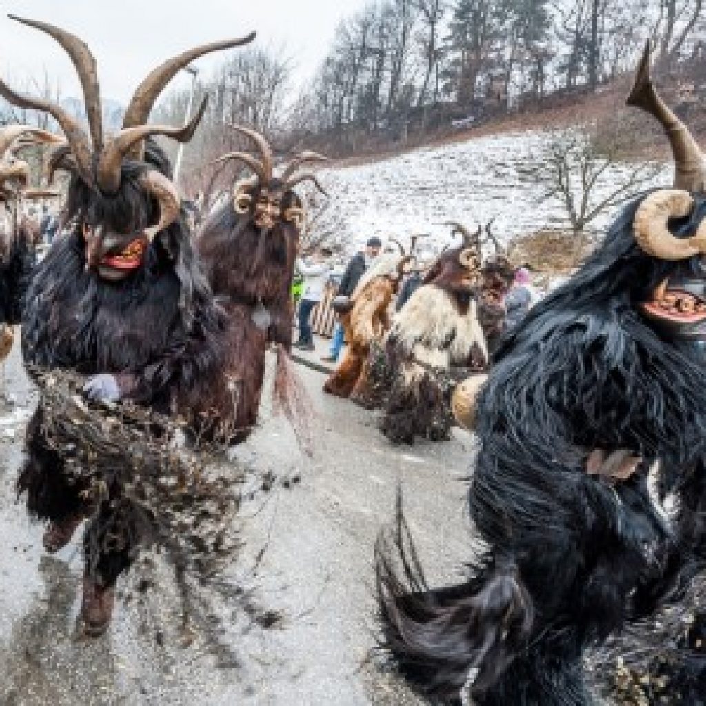 Menschen in pelzigen, gehörnten Kostümen ziehen während eines traditionellen Winterfestes eine verschneite Straße entlang.