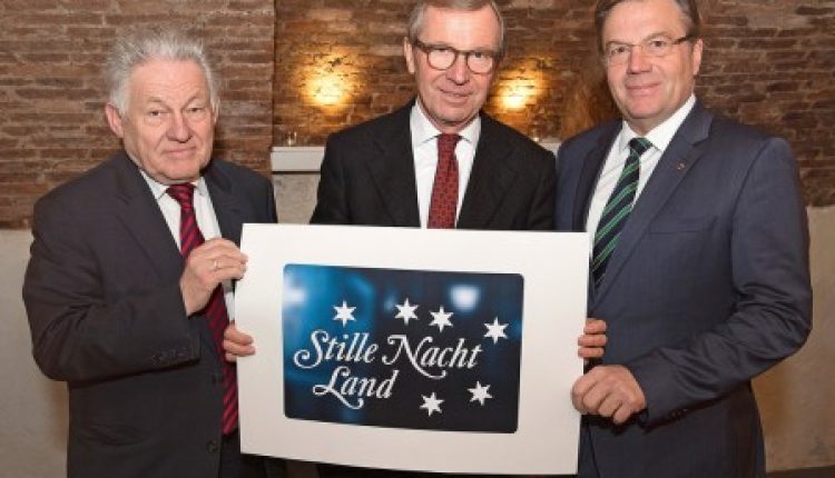 Drei Männer in Anzügen halten ein Schild mit der Aufschrift "Stille Nacht Land" mit Sternen darauf und stehen in einem Haus.