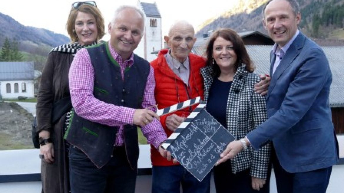 Fünf lächelnde Erwachsene posieren im Freien mit einer Filmklappe; im Hintergrund sind Berge und eine österreichische Kirche zu sehen.