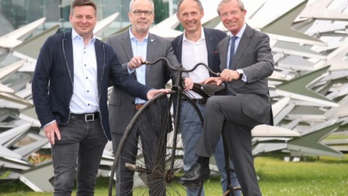Vier Männer in Anzügen lächeln und posieren mit einem altmodischen Fahrrad vor einem modernen Gebäude und einer Skulptur.