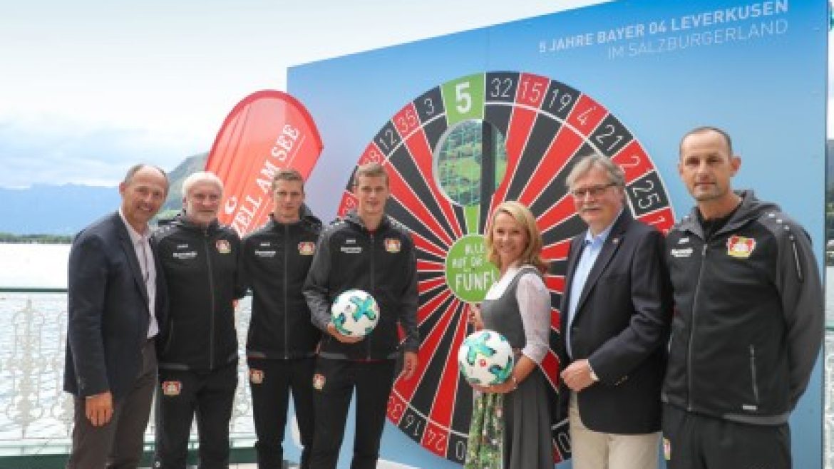 Sechs Personen stehen vor einer großen Dartscheibe, zwei halten Fußbälle, alle lächeln in die Kamera.