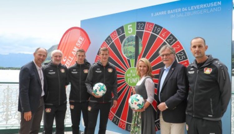Sechs Personen stehen vor einer großen Dartscheibe, zwei halten Fußbälle, alle lächeln in die Kamera.