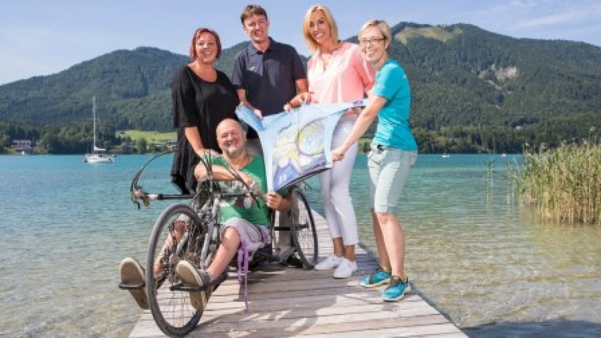 Fünf lächelnde Menschen, einer davon im Rollstuhl, halten eine Landkarte auf einem Steg an einem See mit Bergen im Hintergrund.