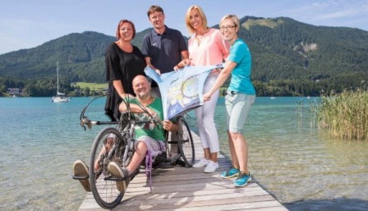 Fünf lächelnde Menschen, einer davon im Rollstuhl, halten eine Landkarte auf einem Steg an einem See mit Bergen im Hintergrund.
