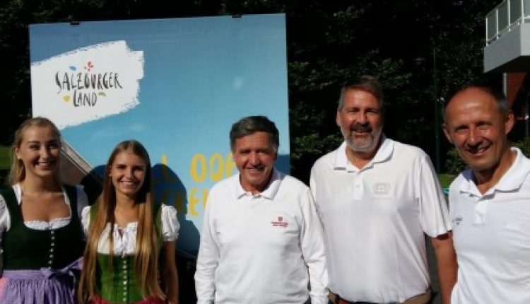 Fünf Personen lächeln im Freien, zwei Frauen in Trachten und drei Männer in weißen Hemden, mit einem Salzburg-Schild.