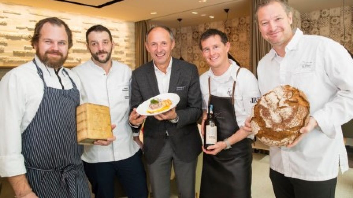 Fünf lächelnde Männer, die Brot, einen Teller und eine Flasche in der Hand halten, stehen in einem Restaurant zusammen.