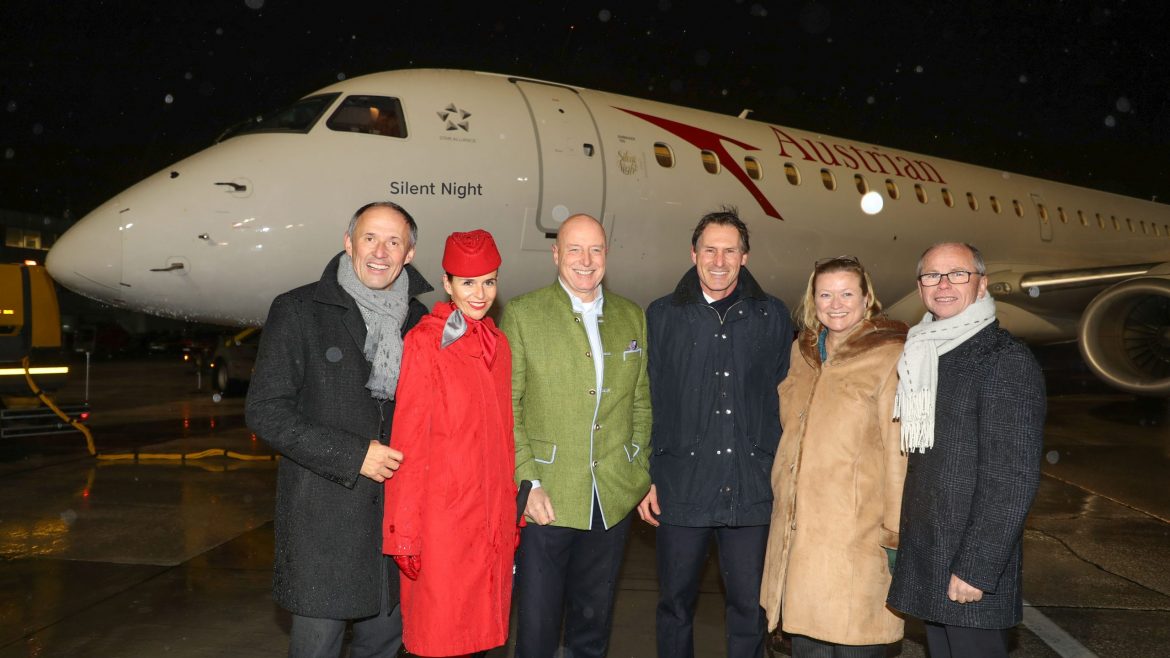 Sechs Personen posieren lächelnd vor einem Flugzeug der Austrian Airlines, das nachts auf einer nassen Landebahn steht.