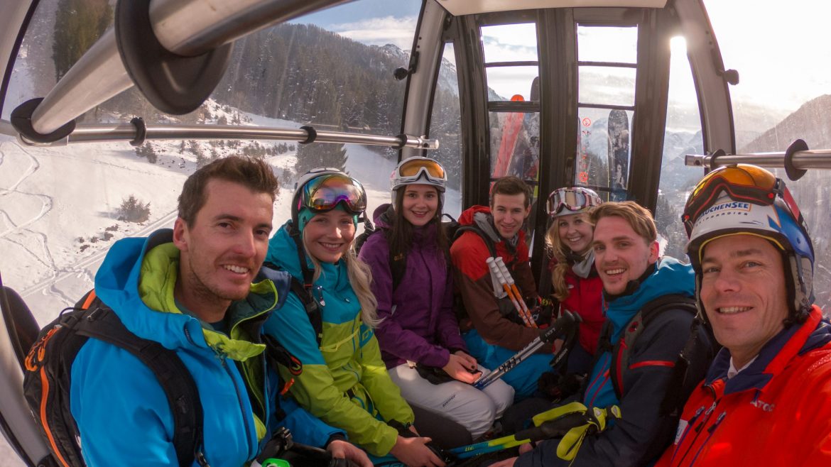 Sieben lächelnde Skifahrer in farbenfroher Kleidung fahren in einer Skigondel, durch deren Fenster verschneite Berge zu sehen sind.