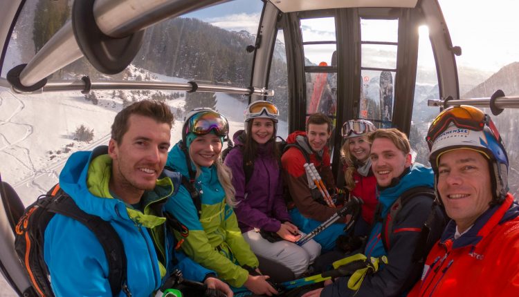 Sieben lächelnde Skifahrer in farbenfroher Kleidung fahren in einer Skigondel, durch deren Fenster verschneite Berge zu sehen sind.