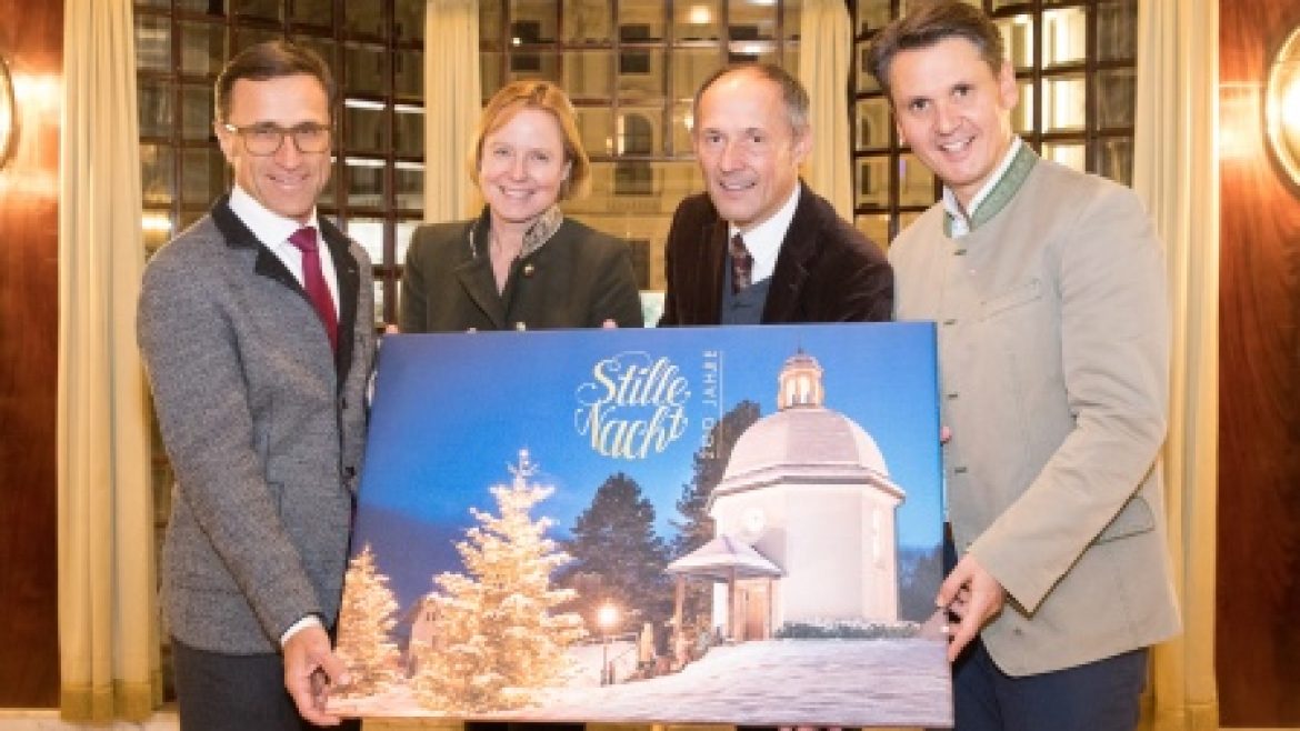 Vier Personen, die lächelnd ein Stille-Nacht-Gemälde einer verschneiten Kapelle mit Weihnachtsbäumen halten.
