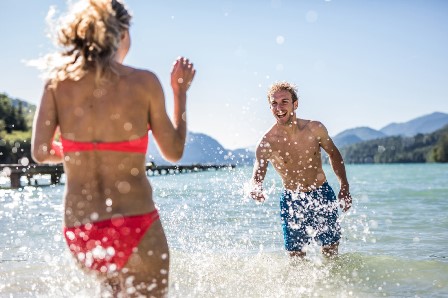 Zwei Menschen spielen und planschen an einem sonnigen Tag im klaren Wasser, während im Hintergrund die Berge zu sehen sind.