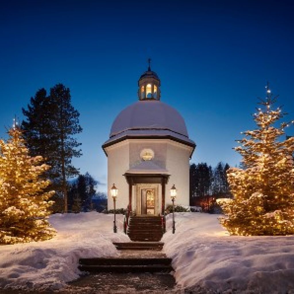 Eine schneebedeckte Kapelle mit beleuchteten Weihnachtsbäumen und Lampen an einem Winterabend.