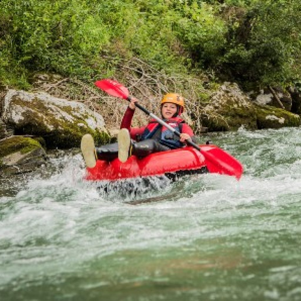 Eine Person in einem roten Kajak paddelt durch einen schnell fließenden Fluss, umgeben von Grün und Felsen.