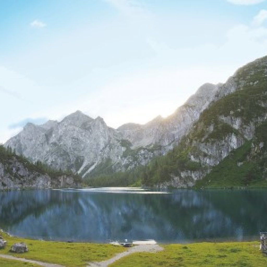 Ein ruhiger See, umgeben von grünen Bergen unter einem klaren Himmel bei Sonnenuntergang, mit Wanderwegen im Vordergrund.