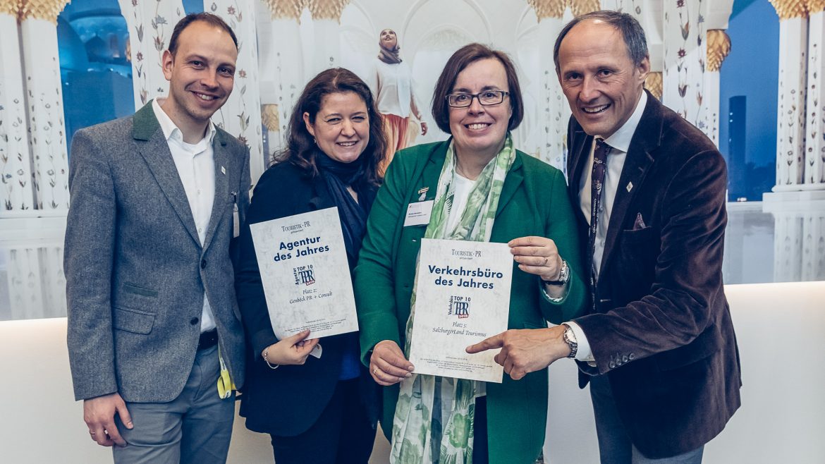 Vier lächelnde Erwachsene halten die Urkunden für die Auszeichnung in der Hand und posieren gemeinsam vor einem dekorativen weiß-goldenen Hintergrund.