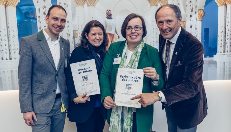 Vier lächelnde Erwachsene halten die Urkunden für die Auszeichnung in der Hand und posieren gemeinsam vor einem dekorativen weiß-goldenen Hintergrund.