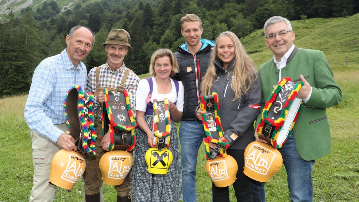 Sechs Menschen in traditioneller Kleidung halten bunte Kuhglocken und lächeln in die Kamera, im Hintergrund grüne Hügel.