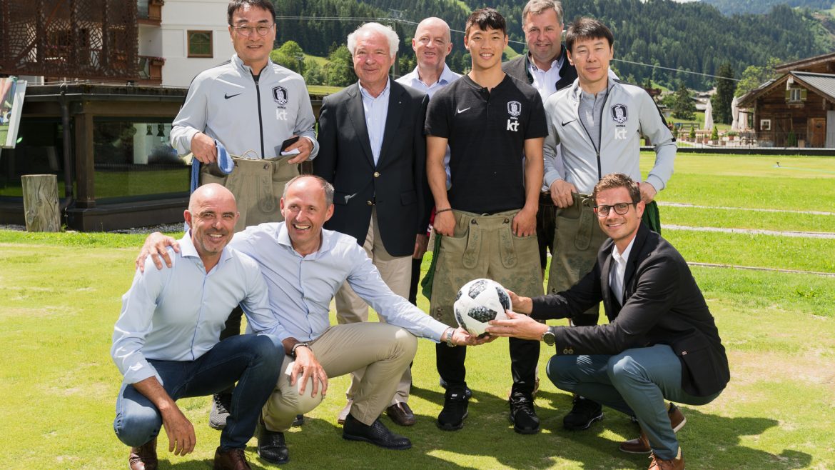 Eine Gruppe von Männern posiert und lächelt im Freien auf einer Wiese, wobei zwei Männer vorne einen Fußball halten.