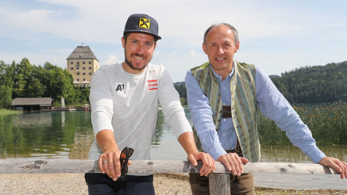 Zwei Männer lächeln an einem See mit einem Gebäude und Bäumen im Hintergrund an einem sonnigen Tag.