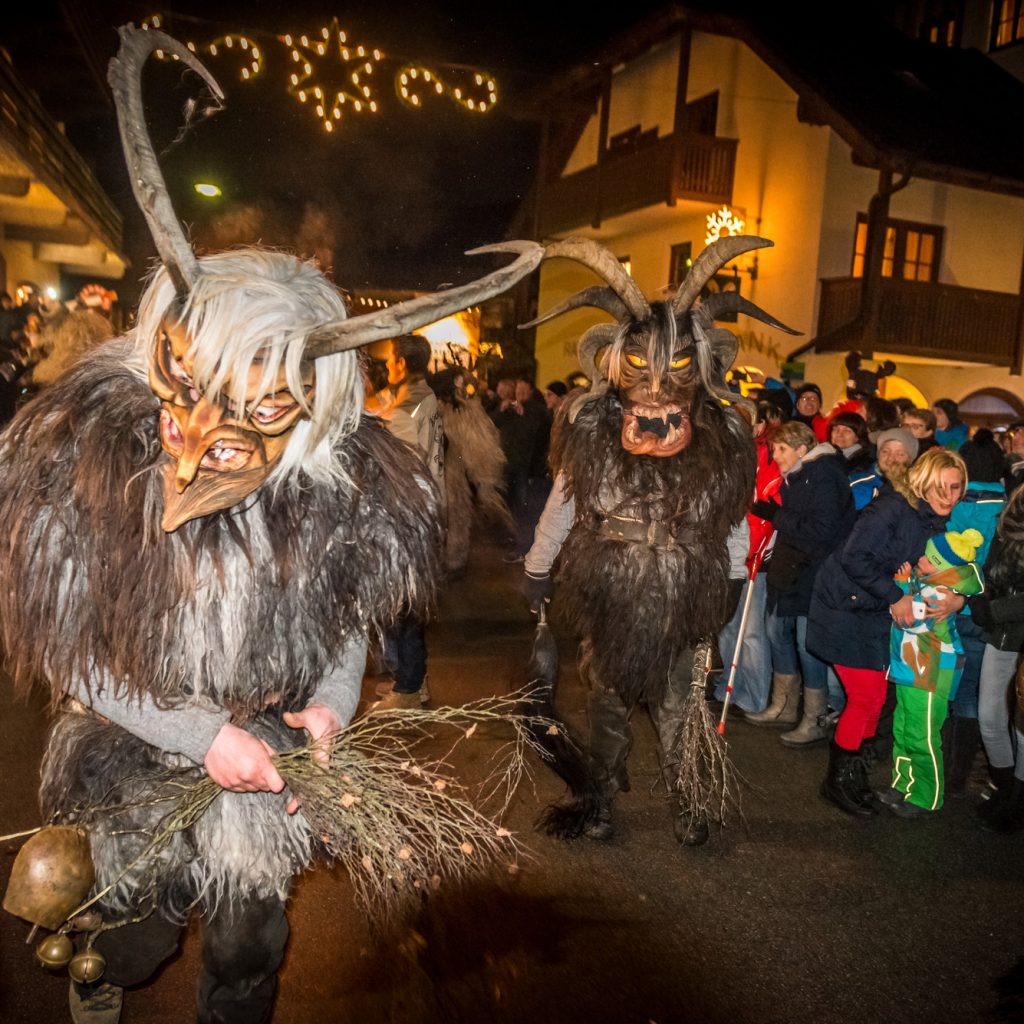 Menschen in Krampuskostümen ziehen nachts während eines Winterfestes durch eine festlich geschmückte, überfüllte Straße.