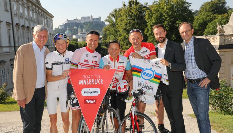 Sieben lächelnde Menschen, einige in Radfahrerkleidung, posieren mit Fahrrädern und einem roten Road to Innsbruck-Banner im Freien.