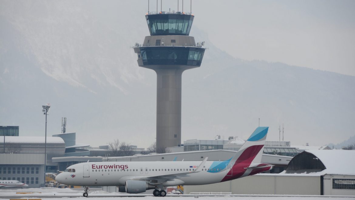 Ein Eurowings-Flugzeug rollt auf einer verschneiten Landebahn mit einem Flughafenkontrollturm im Hintergrund.