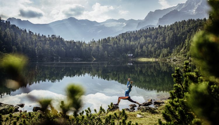 Person beim Yoga an einem ruhigen See, umgeben von Wald und Bergen unter einem teilweise bewölkten Himmel.