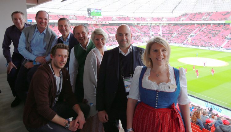 Eine Gruppe lächelnder Menschen in einer Stadionsuite, im Hintergrund ist ein Fußballfeld zu sehen.