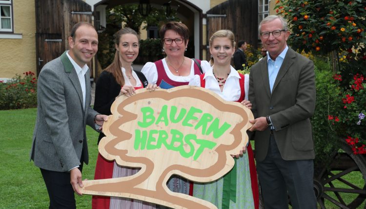 Fünf Personen in traditioneller Kleidung halten ein hölzernes Bauernherbst-Schild und lächeln im Freien neben einem Gebäude und Blumen.