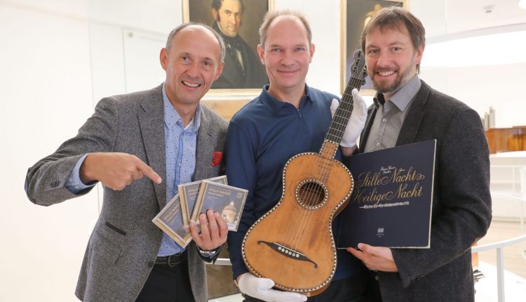 Drei lächelnde Männer posieren mit einer Gitarre, CDs und einer Schallplatte vor gerahmten Porträts.