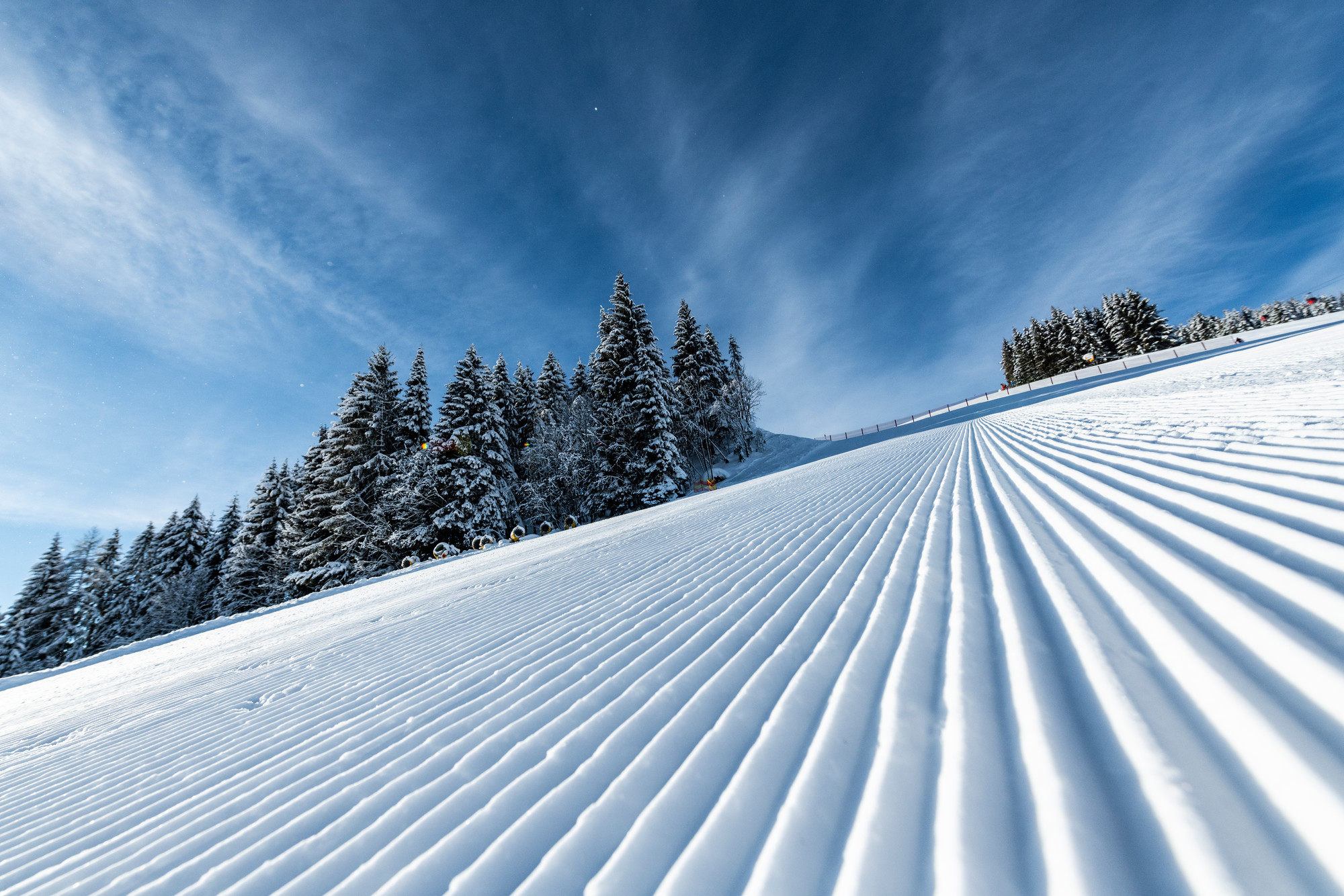 Präparierte Skipiste mit parallelen Linien, verschneiten Bäumen und einem blauen Himmel mit Wolkenfetzen.
