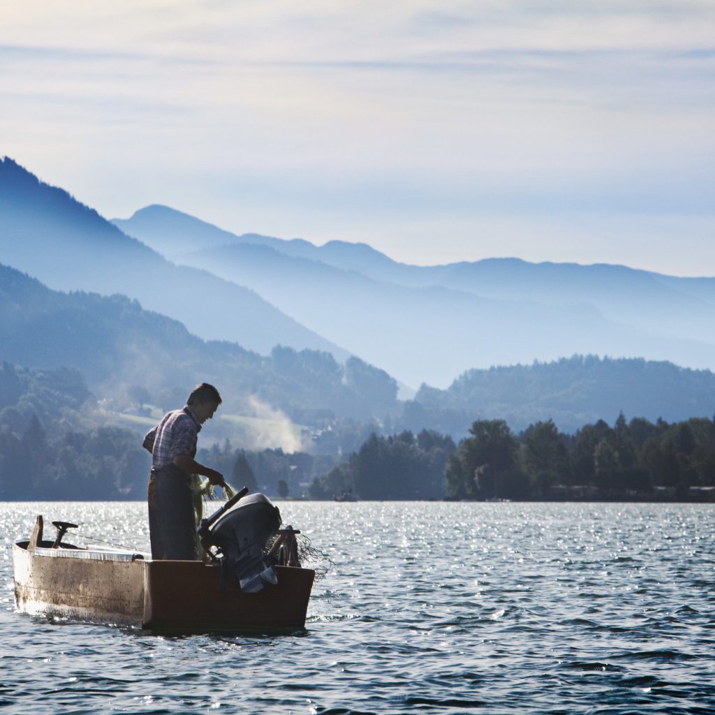 Eine Person steht in einem kleinen Boot auf einem See, umgeben von nebligen Bergen unter blauem Himmel.