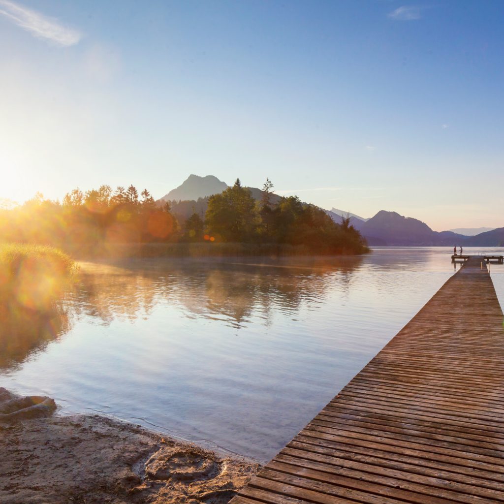 Der hölzerne Steg führt in einen ruhigen See bei Sonnenaufgang, mit Bäumen und Bergen in der Ferne.
