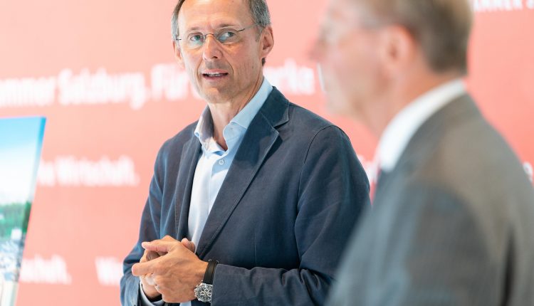 Ein Mann in einem blauen Blazer spricht, während ein anderer Mann in einem Anzug unscharf im Vordergrund steht.