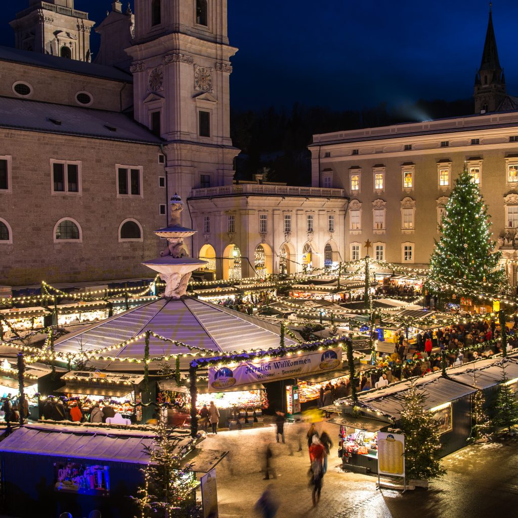 Nächtlicher Weihnachtsmarkt mit festlicher Beleuchtung, einem großen Weihnachtsbaum und Menschen, die zwischen geschmückten Ständen spazieren gehen.