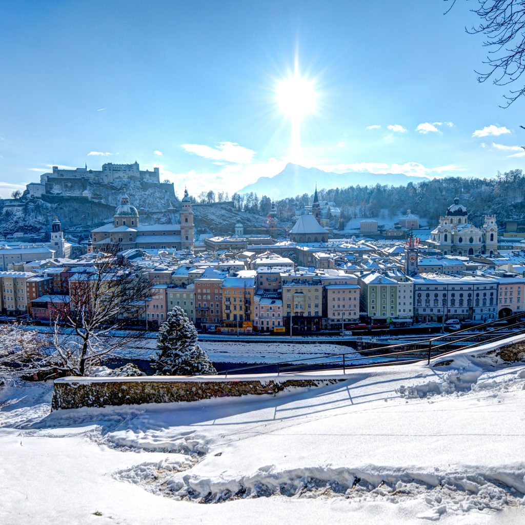 Schneebedeckte Stadtlandschaft von Salzburg, Österreich, mit Sonnenschein und einer Festung auf einem entfernten Hügel.