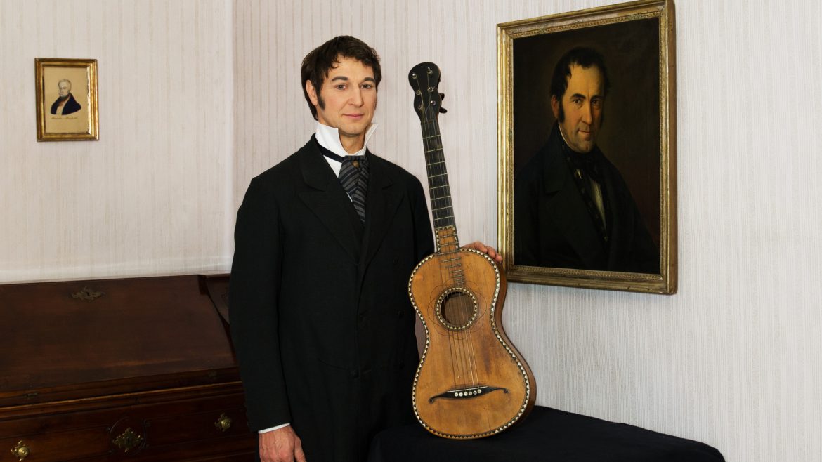Ein Mann in historischer Kleidung steht neben einer antiken Gitarre und einem gerahmten Porträt an der Wand in einem historischen Raum.