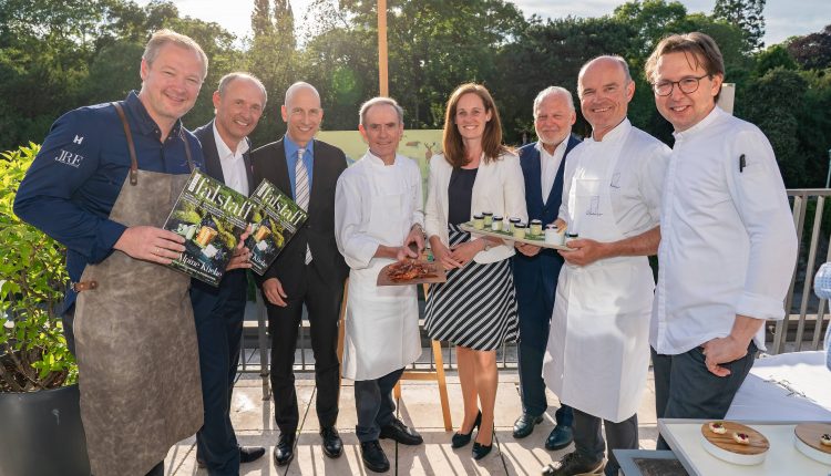 Acht Personen, darunter Köche und Fachleute, posieren an einem sonnigen Tag mit Essen und Zeitschriften im Freien.