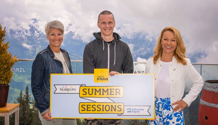 Drei Personen lächeln und halten ein Summer Sessions Schild mit Bergen und bewölktem Himmel im Hintergrund.