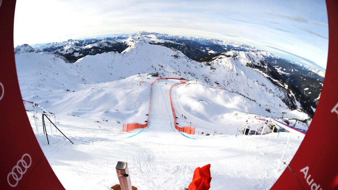 Blick durch einen runden Rahmen auf eine verschneite Skipiste, die sich einen Berg hinunterschlängelt, umgeben von schneebedeckten Gipfeln.
