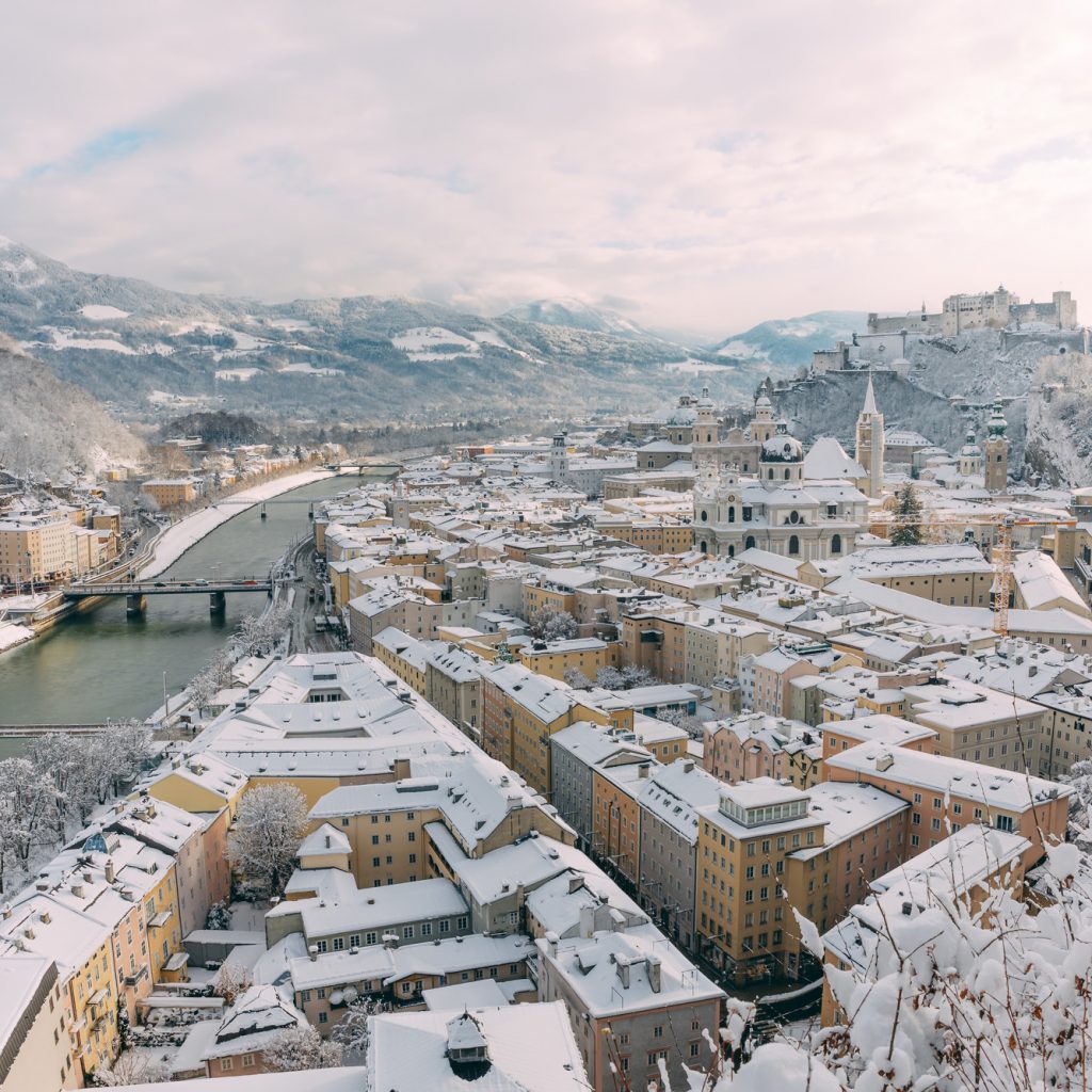 Schneebedeckte Stadtlandschaft mit Flüssen, Brücken und entfernten Bergen unter einem teilweise bewölkten Himmel.