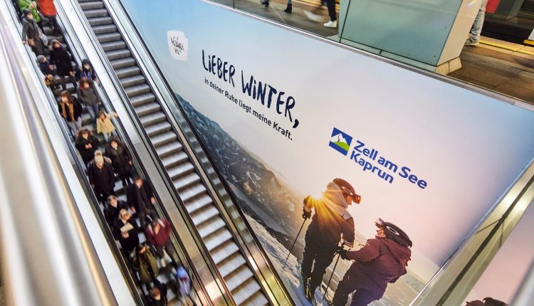 Menschen auf einer Rolltreppe neben einer großen Winterreisewerbung für Zell am See-Kaprun.