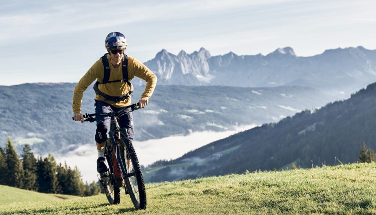 Eine Person fährt mit einem Mountainbike bergauf auf einem grasbewachsenen Hang mit Bergen im Hintergrund.
