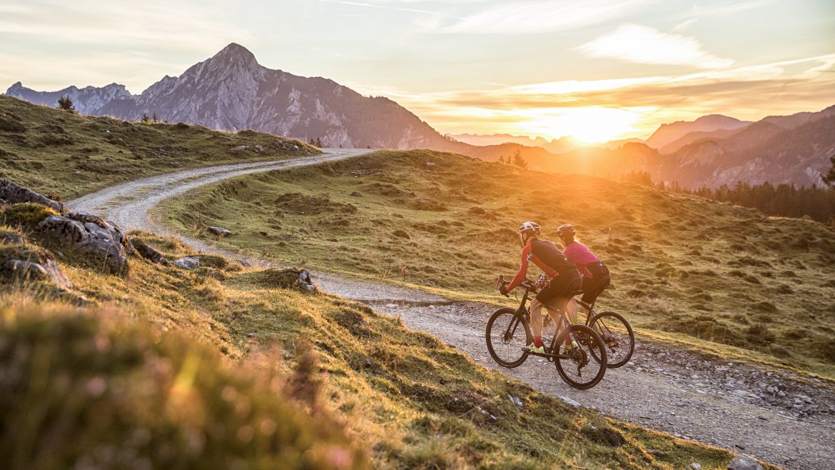 Zwei Radfahrer fahren bei Sonnenuntergang auf einem kurvenreichen Bergweg mit Bergen im Hintergrund.
