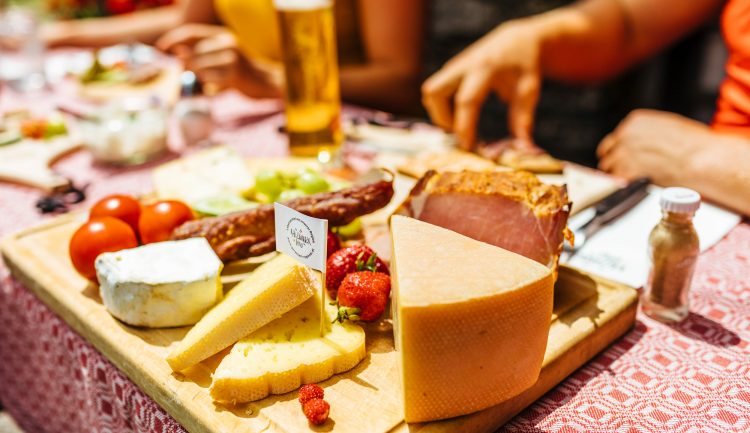 Ein Holzbrett mit verschiedenen Käsesorten, Wurstwaren, Tomaten und Erdbeeren auf einem Tisch im Freien.