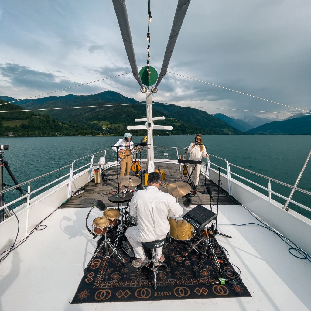 Eine Band tritt auf einem Bootsdeck auf, umgeben von einem See und Bergen unter einem bewölkten Himmel.