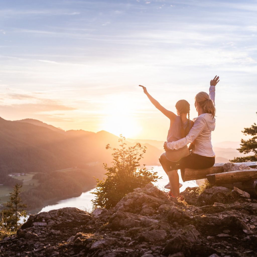 Zwei Menschen sitzen auf einer Bank auf einem Berggipfel und genießen einen malerischen Sonnenaufgang über Hügeln und einem Fluss.