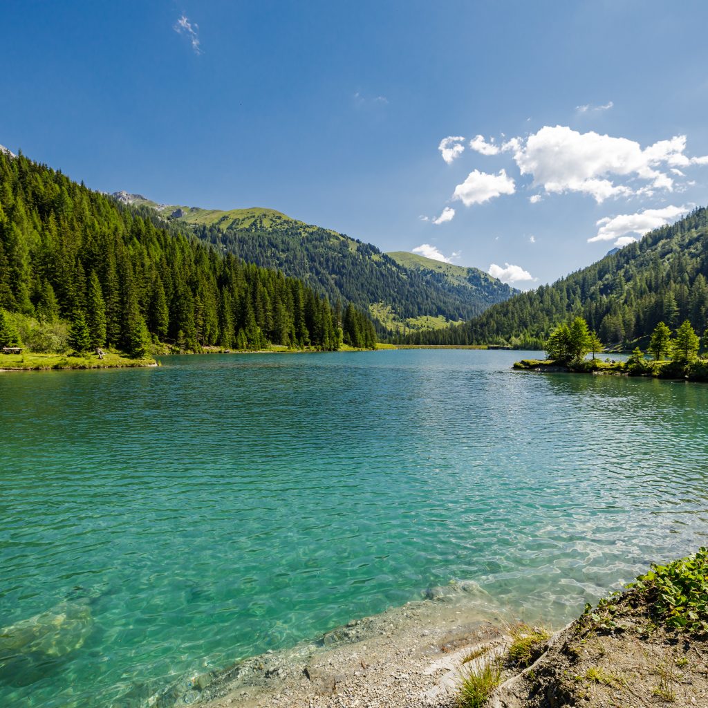 Ein klarer blauer See, umgeben von grünen Kiefernwäldern und Bergen unter einem strahlend blauen Himmel mit vereinzelten Wolken.