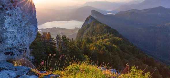 Berglandschaft bei Sonnenaufgang mit Sonnenaufgang, See und üppigen grünen bewaldeten Hügeln in der Ferne.